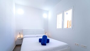A minimalist hotel room with a white bed, two bedside tables, and a window with a view of buildings. - Two bedroom apartment with double beds (TAHICHE)