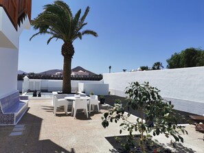 Une terrasse avec des meubles blancs, un palmier et une vue sur une montagne.
