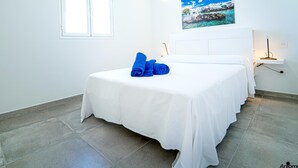 A hotel room with a bed, white walls, a window, a painting of a coastal scene, and a bedside table with a lamp. - Apartment two bedrooms with balcony (TAHICHE)