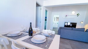 A dining area with a white table set for four, a bottle of wine, and empty wine glasses. There is a grey sofa in the background and a TV mounted on the wall. - Apartment two bedrooms with balcony (TAHICHE)