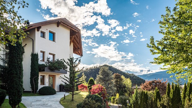 Una casa de dos pisos con un camino de piedra que conduce a la entrada, rodeada de jardines cuidados y un paisaje montañoso de fondo.