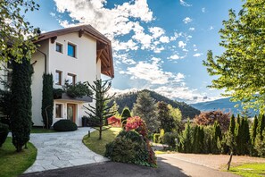 A two-story house with a stone pathway leading to the entrance, surrounded by well-maintained gardens and a mountainous backdrop. - 'Residence Skutial Apartment Santner' with Mountain View, Balconies & Wi-Fi (Castelrotto)