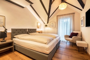 A bedroom with a bed, bedside table, lamp, chair, and a large window with curtains.