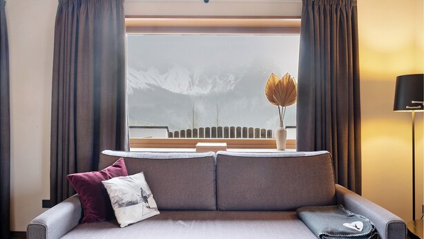 Un soggiorno moderno con un divano grigio, vista sulle montagne attraverso un'ampia finestra e una lampada su un tavolino.