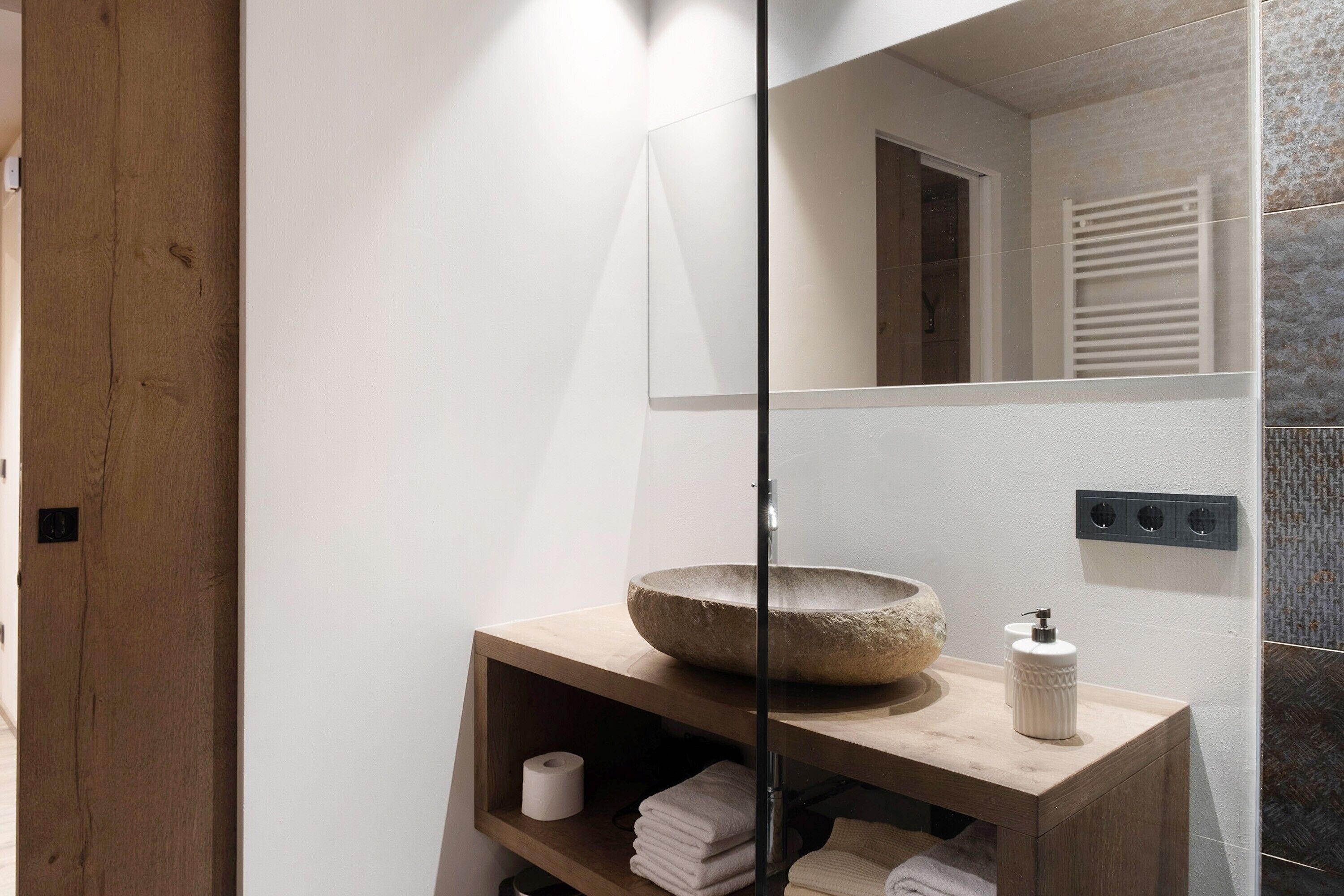 A bathroom with a wooden vanity, a stone basin, a mirror, a towel rack, and a toilet paper roll.