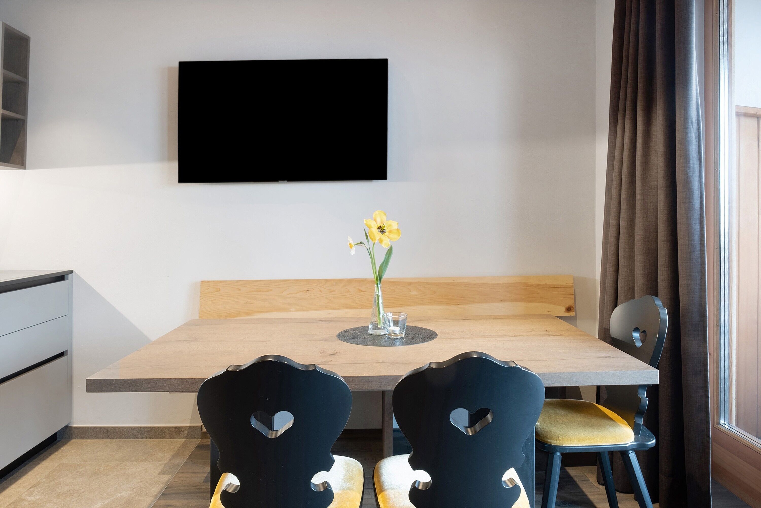 Una habitación moderna con una mesa de comedor de madera, dos sillas, un florero con flores, un televisor de pantalla plana en la pared y una ventana con cortinas.