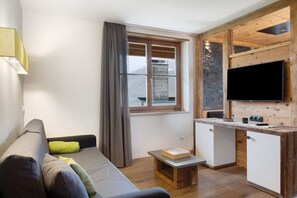 A compact living space with a sofa, a small table, a window with curtains, a kitchen area with a TV, and a stone wall. - Apartment "Angerle Alm Apt Similde B" with Private Terrace, Shared Garden & Wi-Fi (carezza Nova Levante)