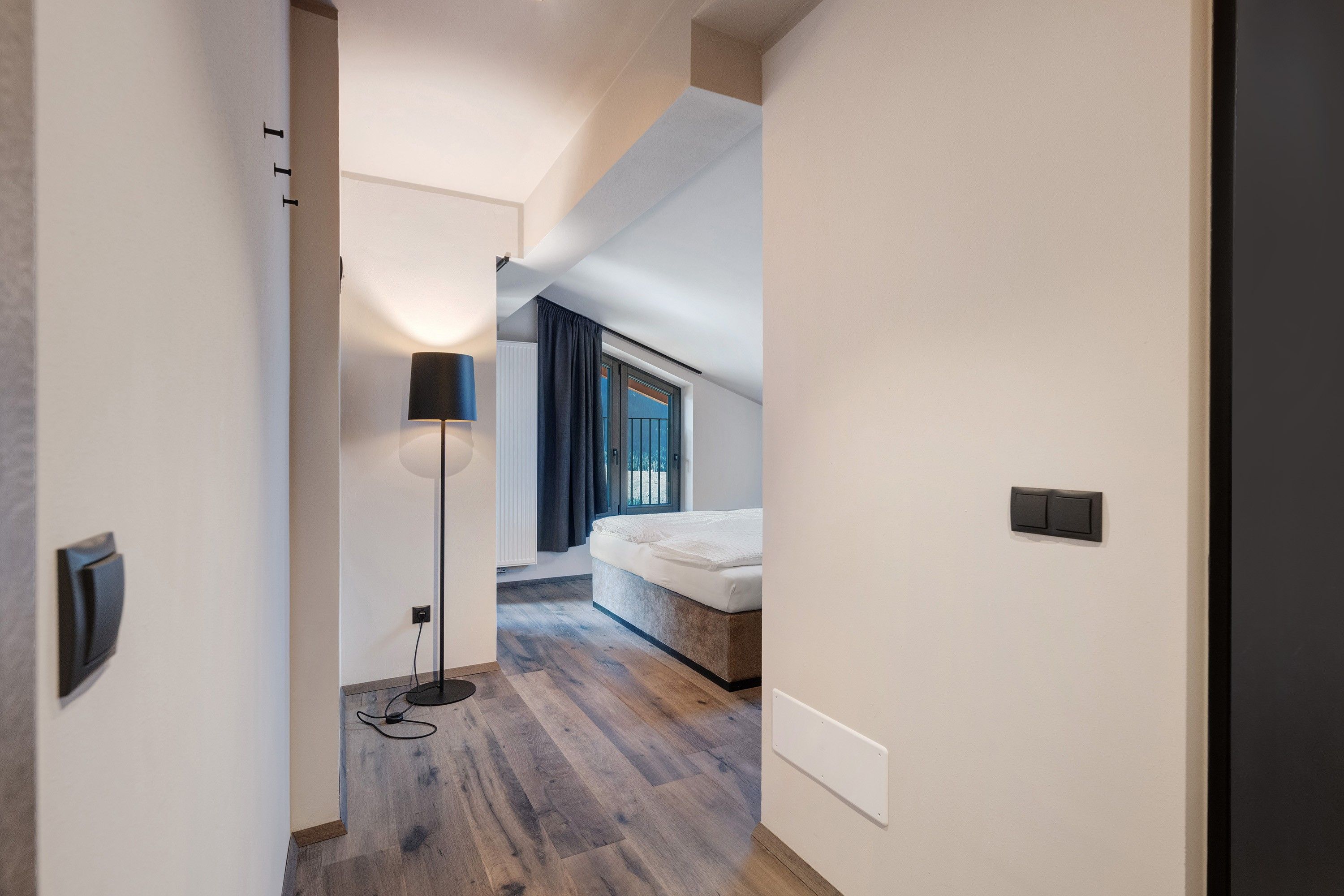 Un couloir étroit mène à une chambre à coucher qui contient un lit, une lampe de sol et une fenêtre avec des rideaux.