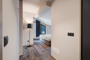 A narrow hallway leading to a bedroom with a bed, a floor lamp, and a window with curtains.