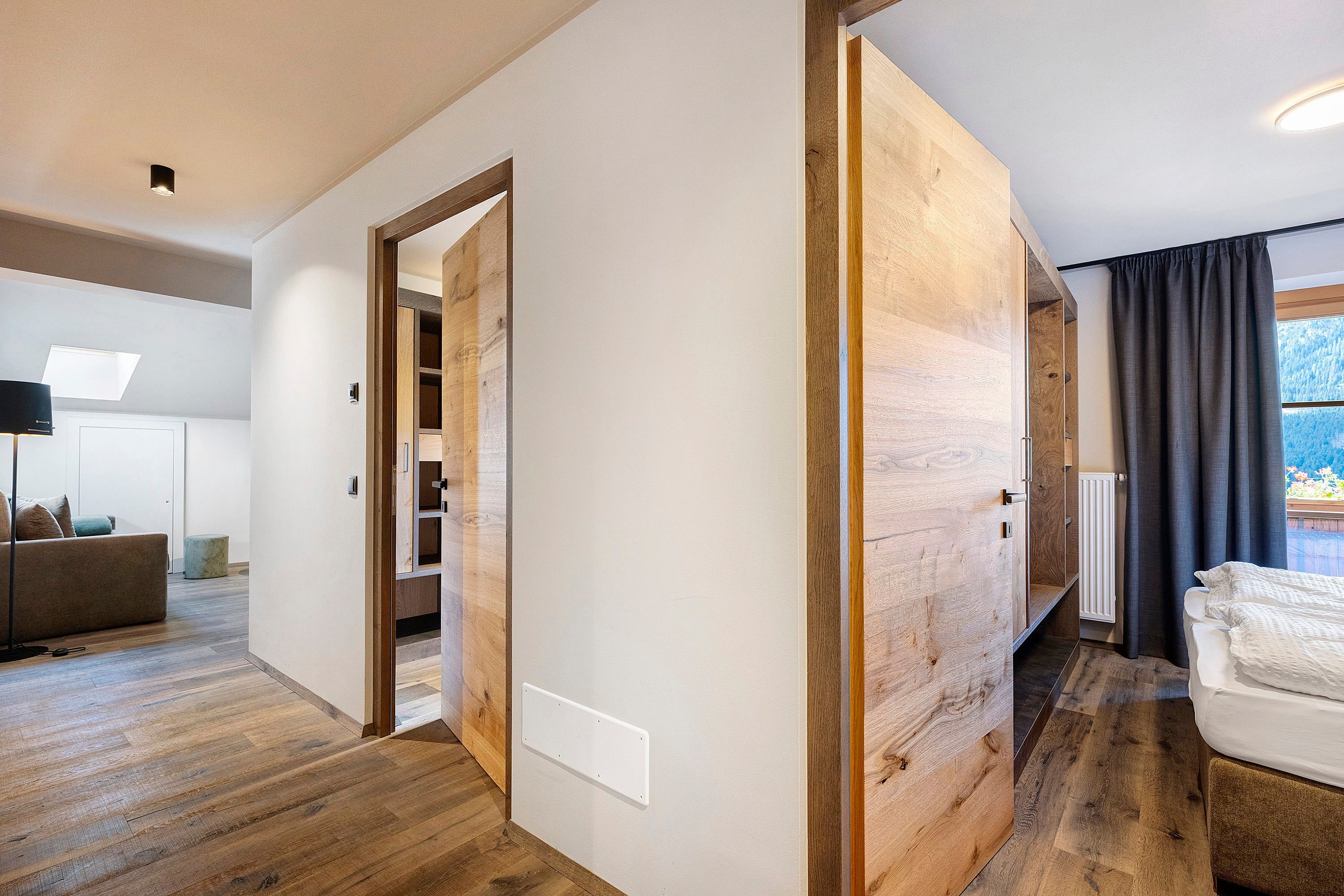 Ein modernes Interieur mit Holzboden, einer weißen Wand, einem Bett mit weißer Bettwäsche und Blick auf eine gebirgige Landschaft durch ein Fenster.