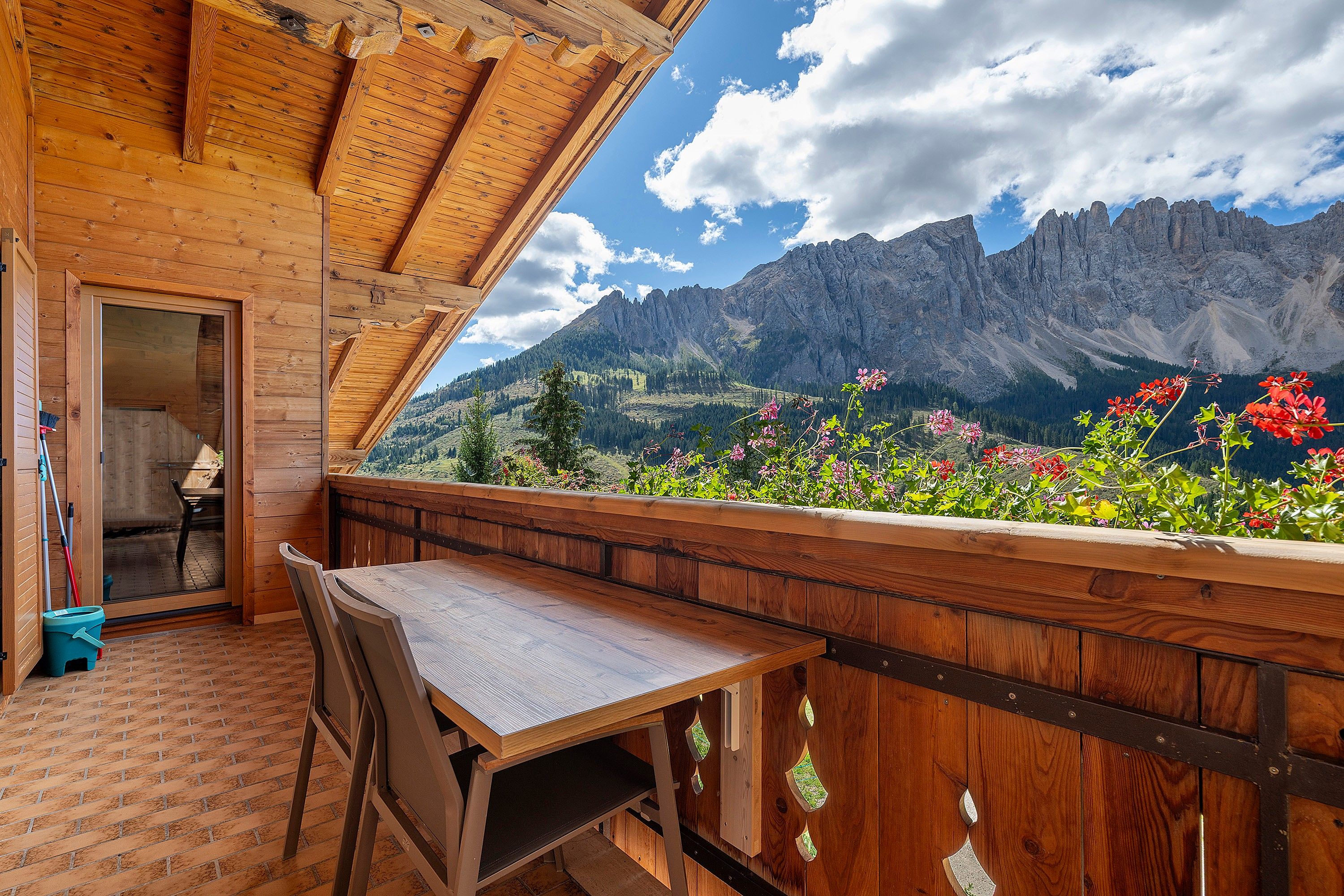 A wooden balcony with a table and chairs, overlooking a mountainous landscape with flowers.