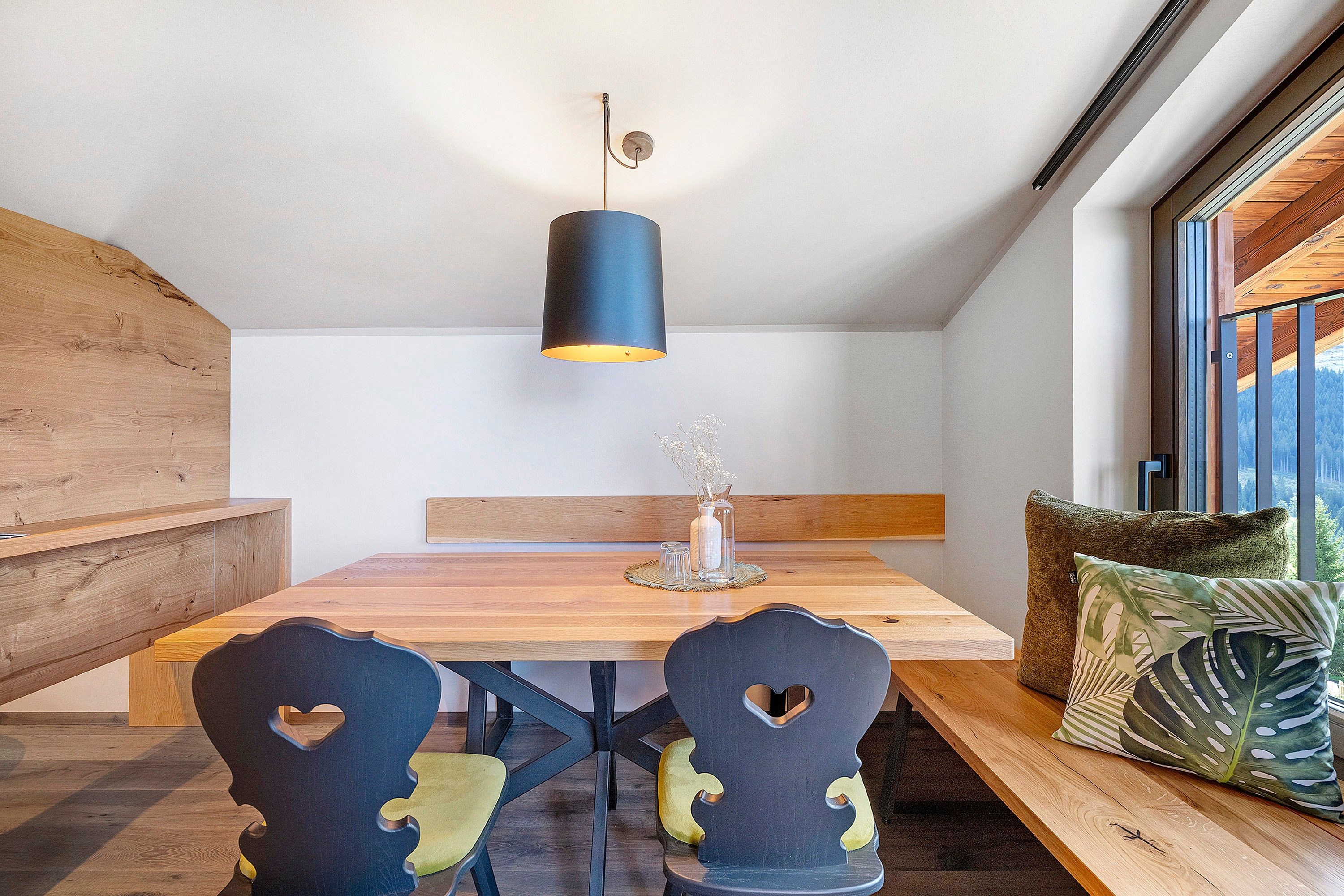 Zona pranzo con tavolo di legno, due sedie con tagli a forma di cuore, una lampada a sospensione e una panca con vista sull'esterno.