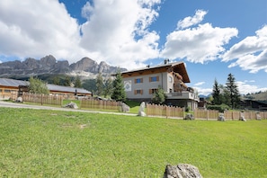 A two-story chalet with a sloped roof, surrounded by a wooden fence, set against a backdrop of mountains and a clear sky.