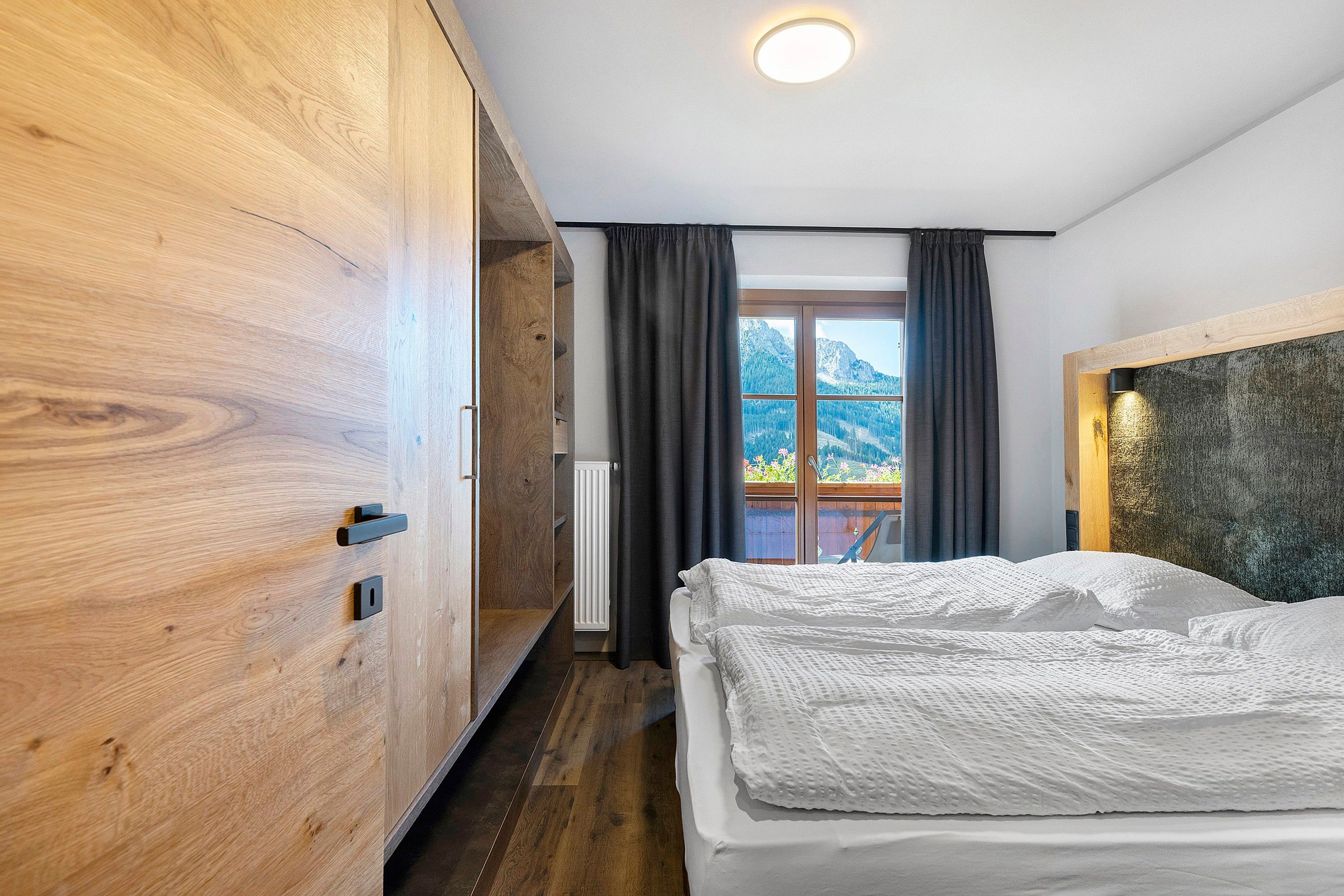 Une chambre avec une armoire en bois, un lit avec une literie blanche, une fenêtre avec des rideaux et une vue sur les montagnes.