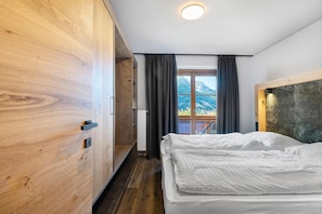 A bedroom with a wooden wardrobe, a bed with white bedding, a window with curtains, and a view of mountains.