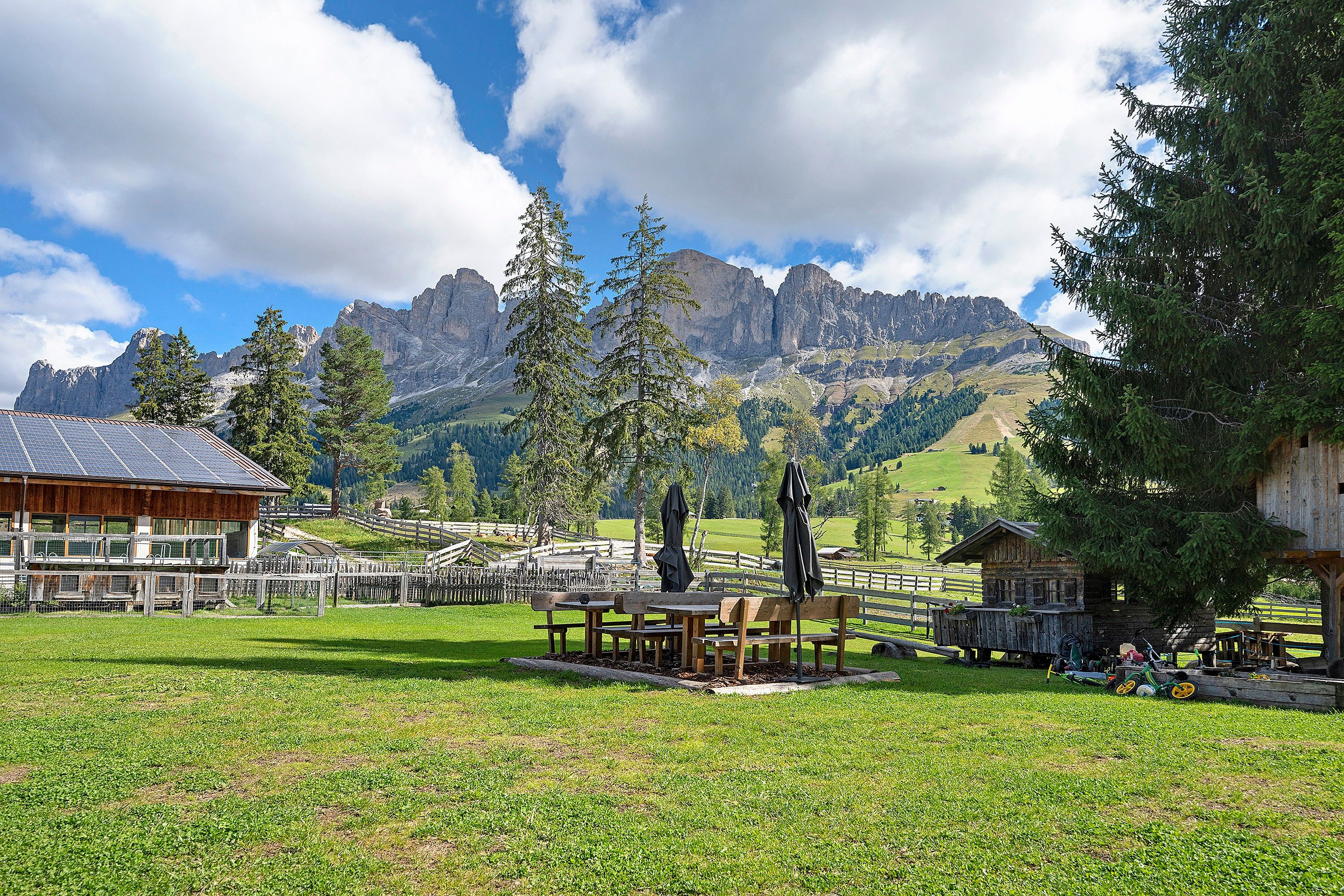 Eine rustikale Berghütte mit Holzbänken und -tischen in einer grünen Wiese, mit Blick auf ein Tal und Berge im Hintergrund.