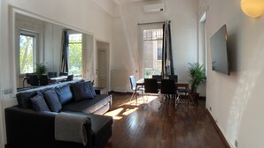 Un salon spacieux avec un canapé de couleur foncée, une table à manger avec des chaises et un téléviseur fixé au mur.