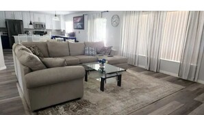 A living room with a beige sectional sofa, a glass coffee table, and a patterned rug. There is a kitchen in the background with white cabinets and a microwave. - The Oaks Bay (Kissimmee)