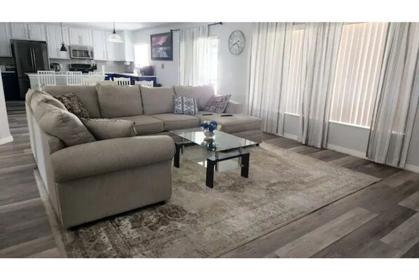 Una sala de estar con un sofá modular color beige, una mesa de centro de vidrio y un tapete estampado. Se observa una cocina al fondo con armarios blancos y un microondas.