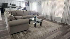 Un salon avec un canapé d’angle beige, une table basse en verre et un tapis à motifs. On aperçoit au fond une cuisine avec des meubles blancs et un four à micro-ondes.