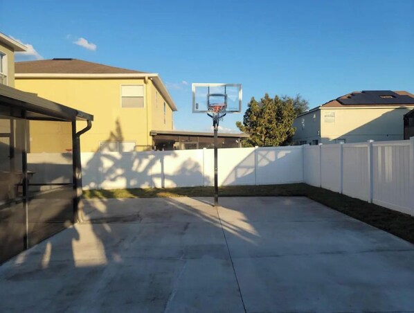 Een achtertuin met een basketbalring, een overdekte patio en een witte hek.