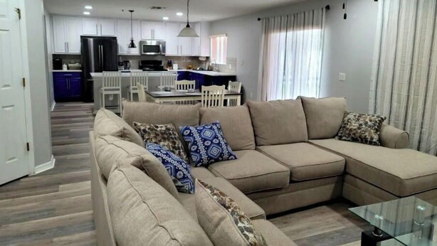 A modern living room with a sectional sofa, a glass coffee table, and a dining area in the background. - The Oaks Bay (Kissimmee)