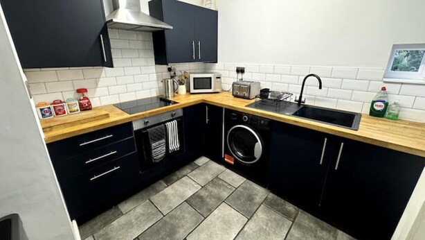 Uma cozinha compacta com armários escuros, uma coifa de aço inoxidável, micro-ondas, pia e máquina de lavar roupa.