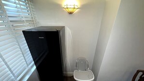 Quarto compacto com geladeira preta, cadeira branca e luminária fixada na parede.
