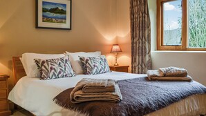 A neatly made bed with a brown blanket and pillows, a bedside lamp, a framed picture on the wall, and a window with curtains. - 3 bedroom accommodation in Keswick (Keswick)