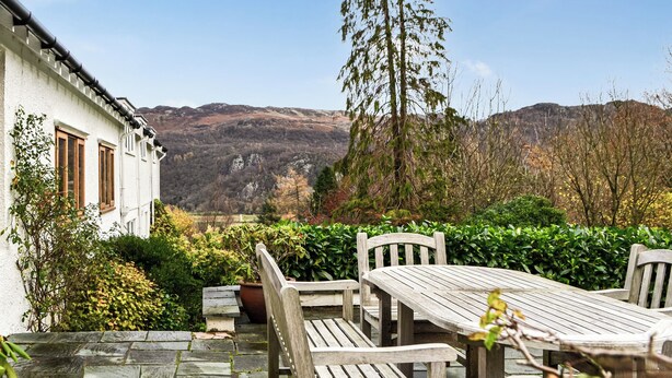 A stone patio with a wooden table and chairs, surrounded by greenery and a view of hills. - 3 bedroom accommodation in Keswick (Keswick)