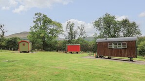 Um cenário rural com uma casa de banho rústica verde e vermelha, uma caravana vermelha e um celeiro de madeira.