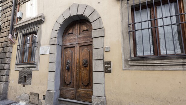 Uma porta de madeira tradicional com um arco de pedra, uma janela com grades de metal e uma pequena placa na parede.