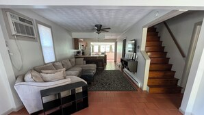 Un salon comprenant un canapé d’angle, une table basse noire, un tapis à motifs, un ventilateur de plafond et un escalier menant à un autre niveau.