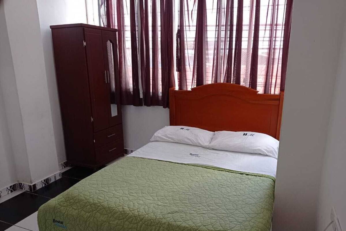 A single bed with a green quilt, a wooden wardrobe, and a window with sheer curtains.