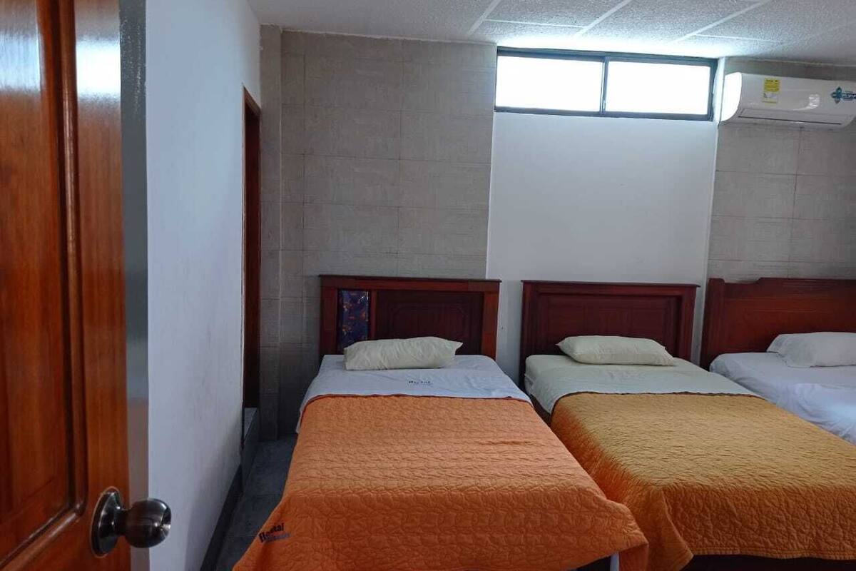 A room with three single beds, each with a wooden headboard and a white mattress. There is a window above the beds, and an air conditioning unit mounted on the wall.