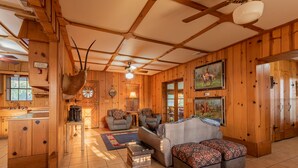 A wooden cabin interior with a sofa, ottoman, ceiling fan, and a mounted antler. - Authentic Ranch Home on 650 Acres with Peace and Tranquility (Marble Falls)