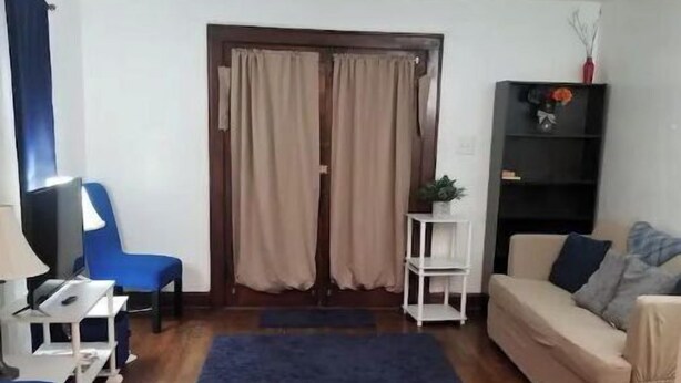 A living room with a wooden floor, a blue rug, a beige sofa, a blue chair, a small white table, a black bookshelf, and a ceiling fan. - Cozy Emerson Heights Bungalow (Indianapolis)