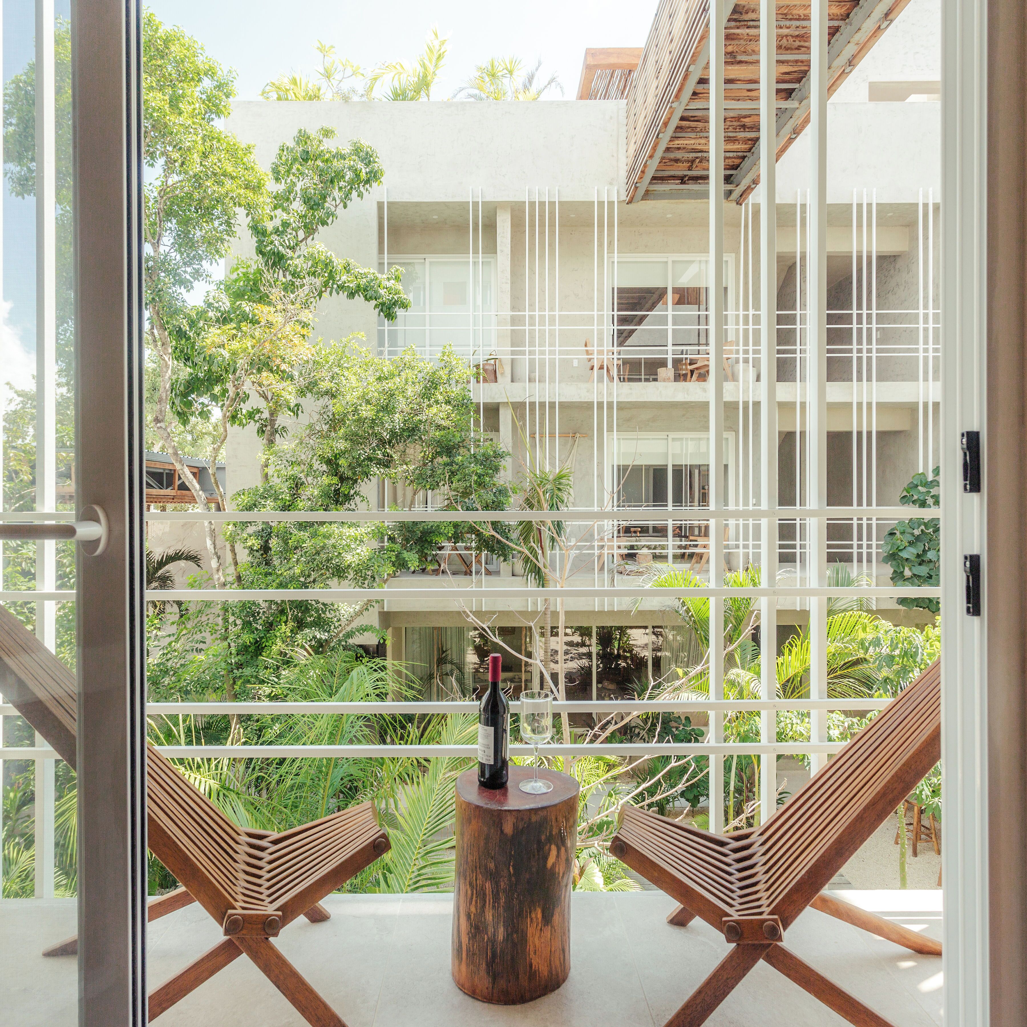 A balcony with two wooden lounge chairs and a small wooden table, overlooking a modern building with multiple balconies and lush greenery.