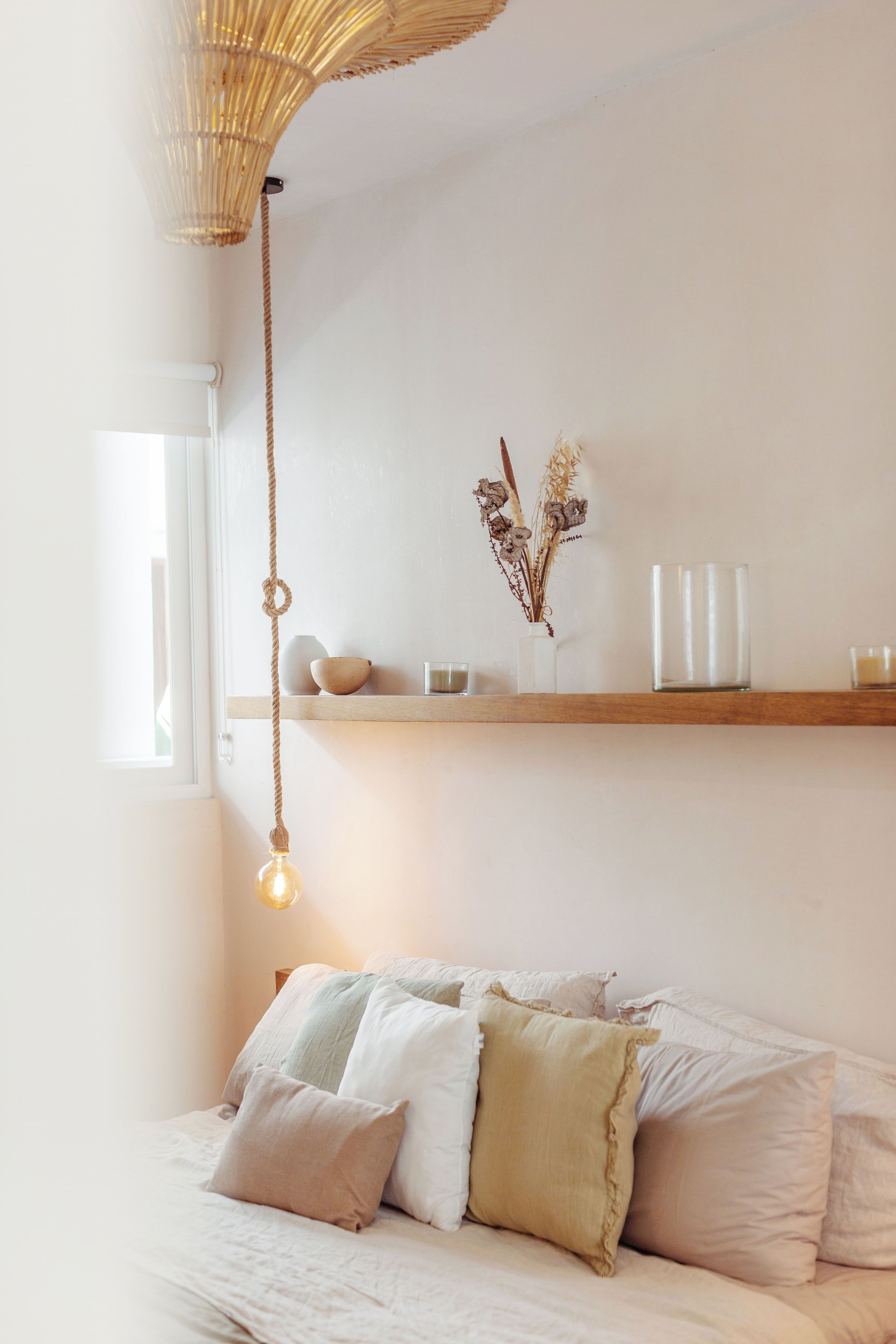 A bedroom with a bed, pillows, a hanging light fixture, a shelf with decorative items, and a window.
