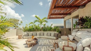 Un espacio exterior moderno con un sofá curvo, mesas redondas de madera y una jardinera de piedra con tuberías de cobre.