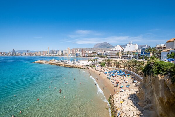A coastal cityscape with a beach, clear blue waters, and a rocky shoreline.