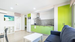 A modern living room with a grey sofa, a white coffee table, and a dining area with a white table and chairs. A kitchen with green cabinets and a stainless steel range hood is visible in the background. - La Rosita Old Town Apartments B-19 (Benidorm)