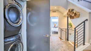 A laundry room with a front-loading washer and dryer, a hallway leading to a bedroom with a bed and a nightstand, and a staircase with a wooden railing. - City Skyline Views: Upscale Denver Townhome (Denver)