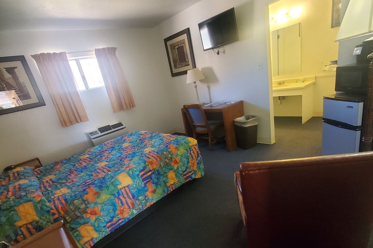 A hotel room with a bed, a desk, a chair, a TV, a small fridge, and a bathroom visible through an open door.