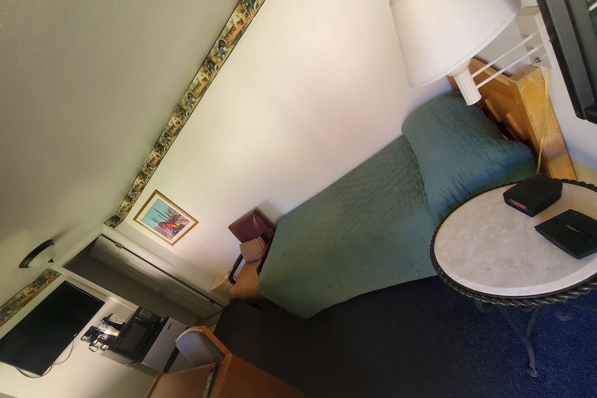 A room with a bed, a small table, a chair, a desk with a computer, and a wall-mounted shelf.