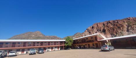A motel with a dirt parking lot, a building with multiple rooms, and a mountainous backdrop.