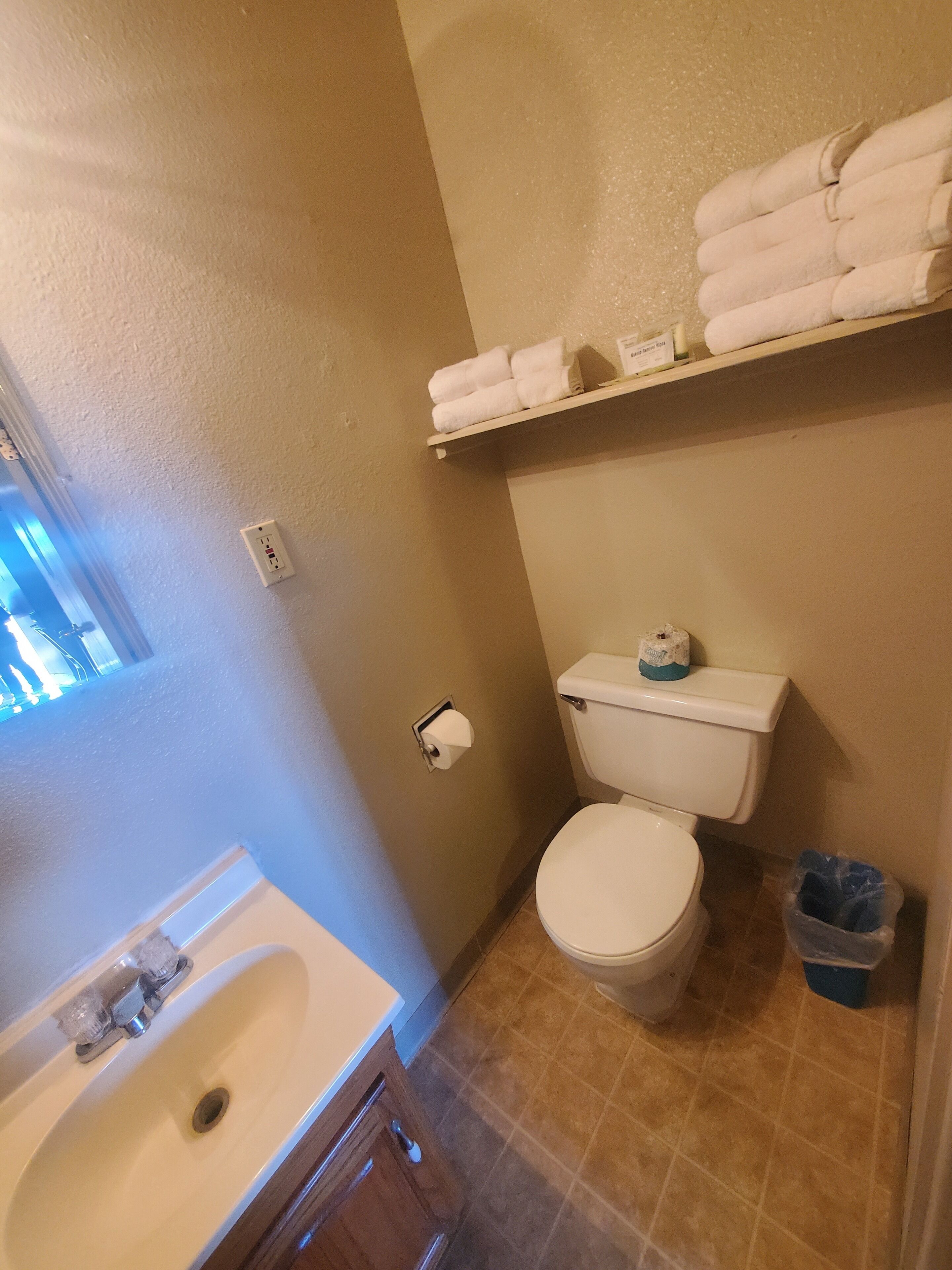 A bathroom with a white sink, a toilet, a shelf with folded towels, and a trash bin.