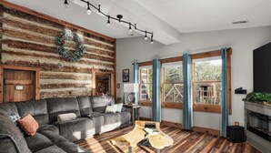 Ein gemütliches Wohnzimmer mit einem Ecksofa, einem Holztisch, einem Kamin und großen Fenstern mit Vorhängen.