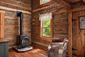 A log cabin interior with a wood-burning stove, a window with a view of greenery, and a patchwork chair.