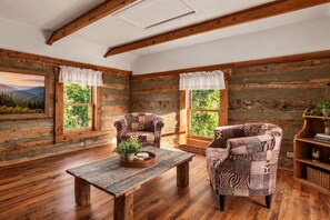 A rustic cabin interior with wooden walls, two upholstered chairs, a wooden table with a plant, and a bookshelf.